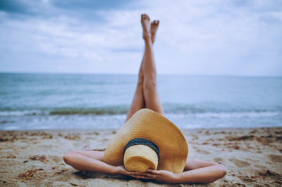 Cerix: En person ligger afslappet på maven på en sandstrand med en stor stråhat over ansigtet og benene løftet op mod himlen. Billedet fremhæver rumpe og ben og kan bruges til at illustrere behandlinger som BBL (Brazilian Butt Lift) hos Cerix.