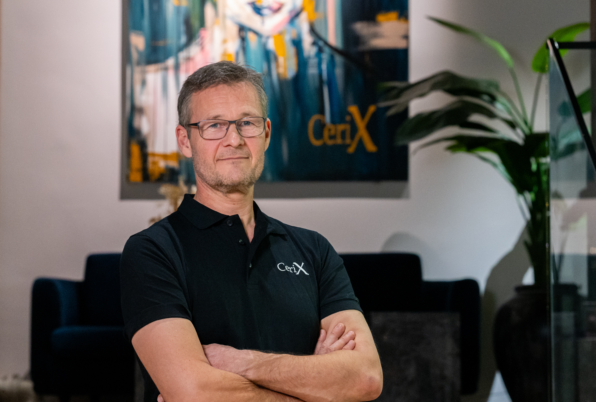 En mand iført Cerix-poloshirt står med korslagte arme i en kliniklobby, Cerix-logo ses i baggrunden. Billedet illustrerer Cerix som virksomhed og viser fokus på ansigt og overkrop, fx ansigtsbehandling såsom Botox, i en rolig, professionel atmosfære.