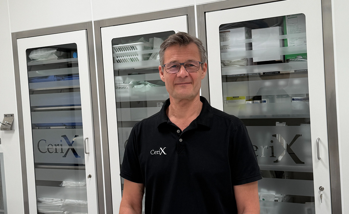 Her er en ALT-tekst til billedet: Billede af en plastikkirurg fra Cerix, der står i et behandlingsrum foran skabe med medicinsk udstyr. Han bærer en sort polo med Cerix-logo. Klinikken er specialiseret i kosmetiske behandlinger som brystforstørrende operation (BFO), Brazilian Butt Lift (BBL) og Botox til ansigtet.