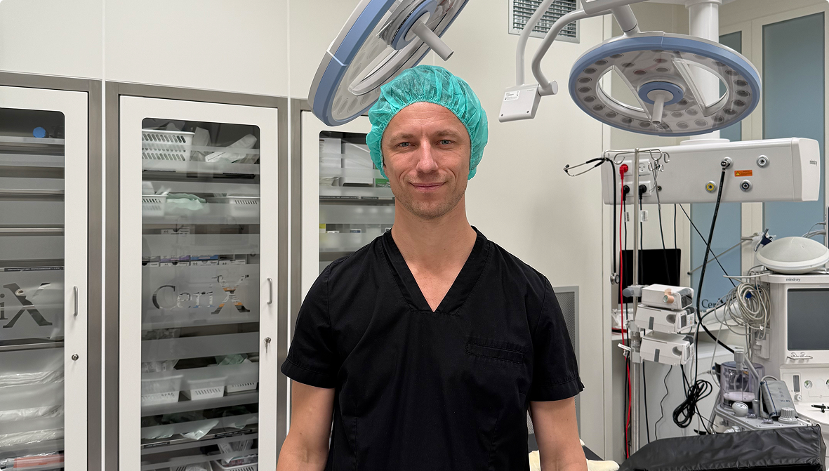 En smilende kirurg står klar i en steril operationsstue hos Cerix. Han bærer sort tøj og en grøn hue, omgivet af avanceret hospitalsudstyr og skabe med Cerix-logo. Her udføres større kosmetiske indgreb på kroppen, såsom brystforstørrende operationer.