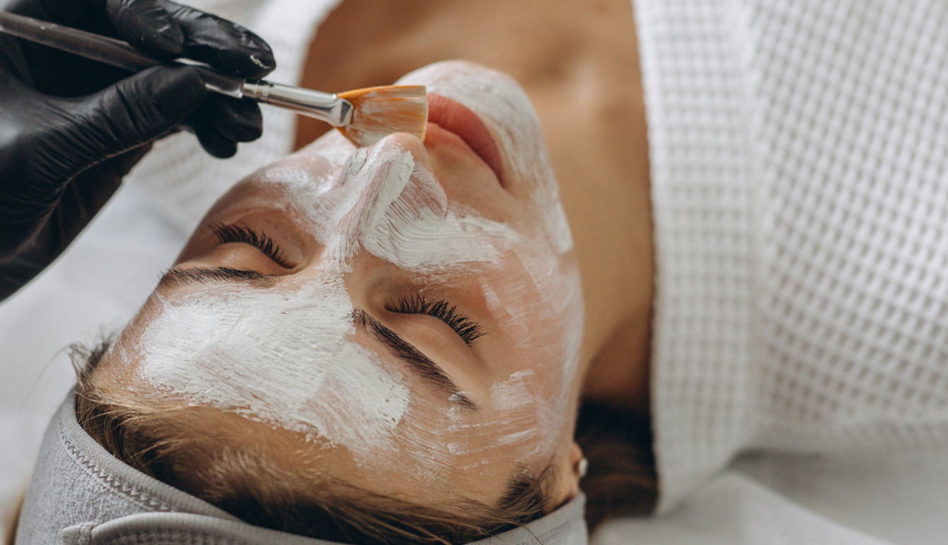 Her er et forslag til ALT-teksten: Et nærbillede af en kvinde, der modtager en professionel ansigtsbehandling hos Cerix. En behandler iført sorte handsker anvender en pensel til omhyggeligt at påføre en hvid maske eller peeling i hele ansigtet. Billedet illustrerer en hudplejeprocedure med fokus på forbedring af huden i ansigtet.