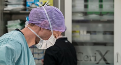 En kirurg iført operationsdragt, maske og lilla operationshætte arbejder i en operationsstue. Billedet illustrerer Cerix som virksomhed inden for kosmetiske og plastikkirurgiske behandlinger, eksempelvis kirurgi på kroppen. Den konkrete behandling og kropsdel, såsom bryster, mave eller ansigt, fremgår ikke af billedet.