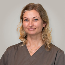 Portræt af smilende voksen kvinde med bølget, skulderlangt hår, mørk uniform og smykker mod neutral baggrund.