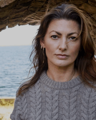 Cerix billede af en kvinde med brunt hår i en striktrøje, der står udendørs ved havet under en stenbue. Fokus er på hendes ansigt, som kan have modtaget en kosmetisk ansigtsbehandling som Botox eller filler hos Cerix for et naturligt og velplejet look.